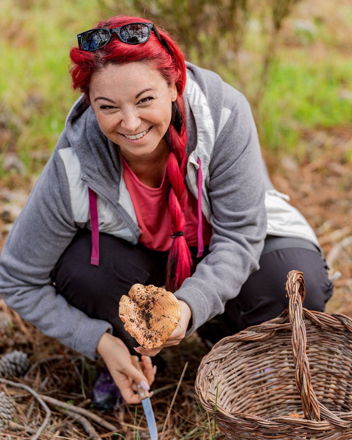 A red haired woman foraging for mushrooms in a forest with a wicker basket. holding a large mushroom with dirt on it.